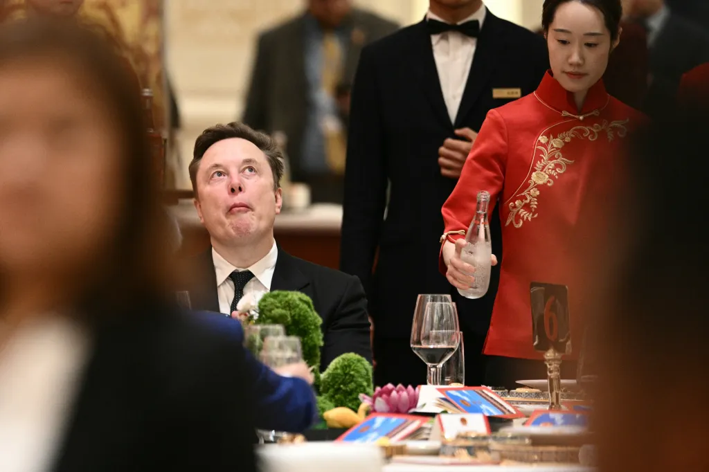 Elon Musk looks up while seated at a banquet table as a server in a red uniform pours water.