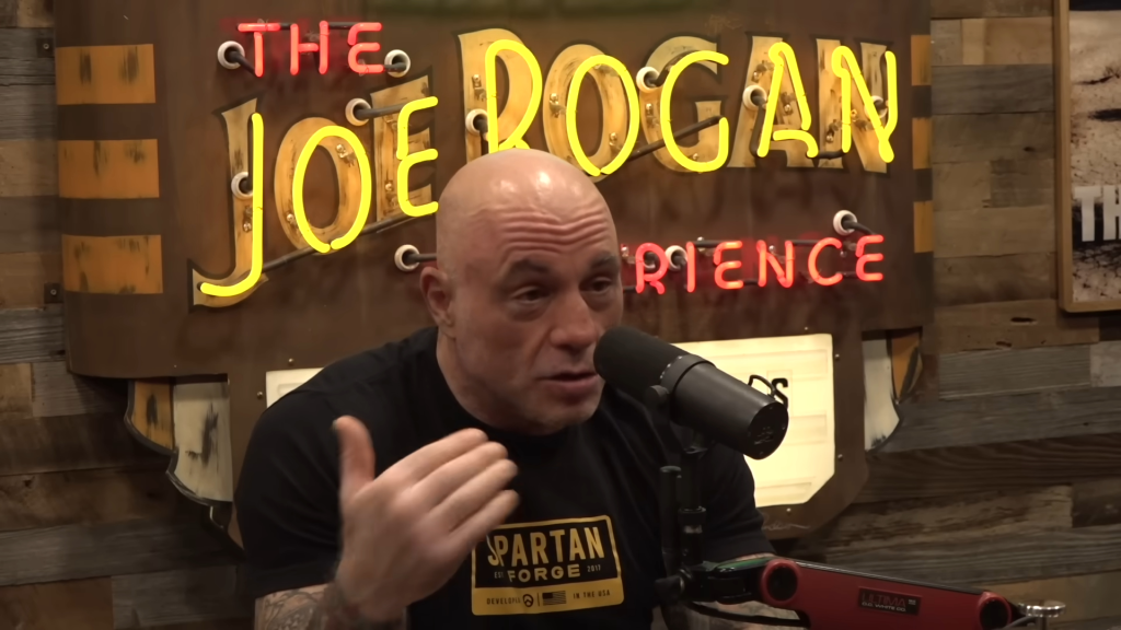 Joe Rogan speaking into a microphone with a neon "The Joe Rogan Experience" sign in the background.