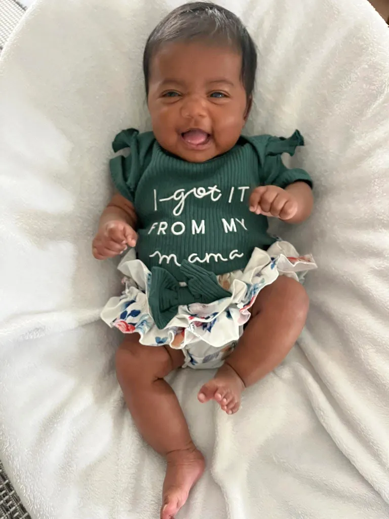 Six-month-old baby Shea smiling while lying on a white blanket, wearing a green bodysuit that says "I-got IT FROM MY mama".