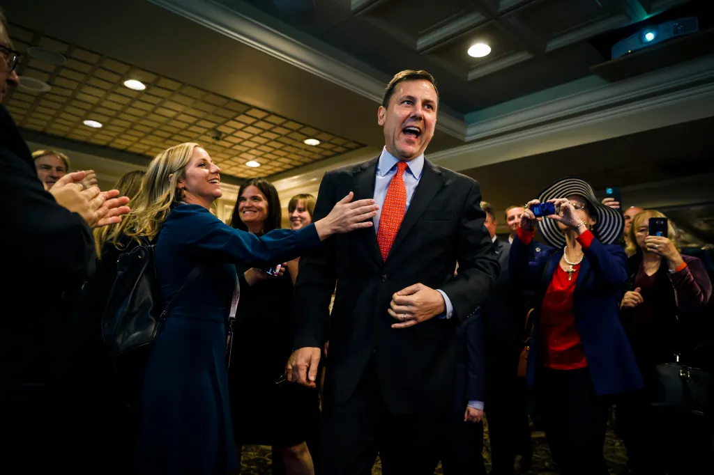 Tom Kean Jr., wearing a suit and red tie, smiles and raises his hand, surrounded by supporters, one of whom has her hand on his arm and others who are clapping and taking photos.