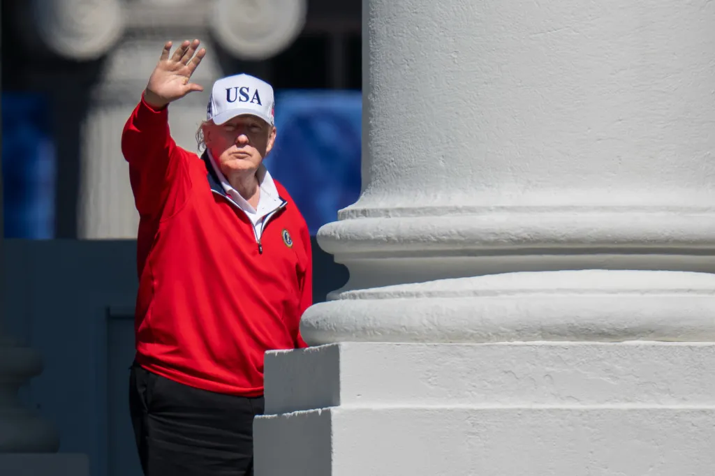 Donald Trump in a red shirt and a white "USA" hat waves as he returns from his golf course.