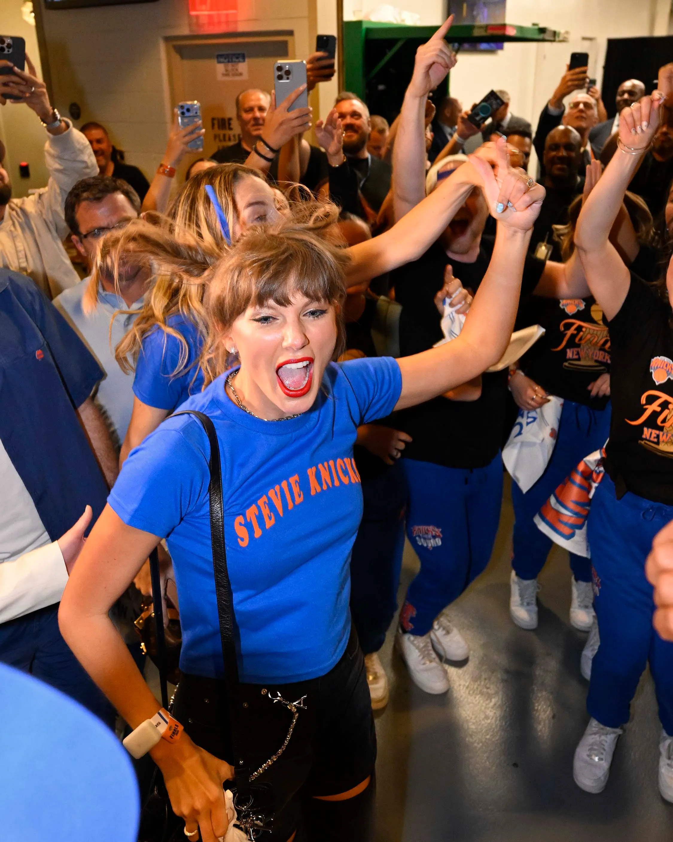 Taylor Swift in a blue "Stevie Knicks" t-shirt, celebrating at a basketball game.