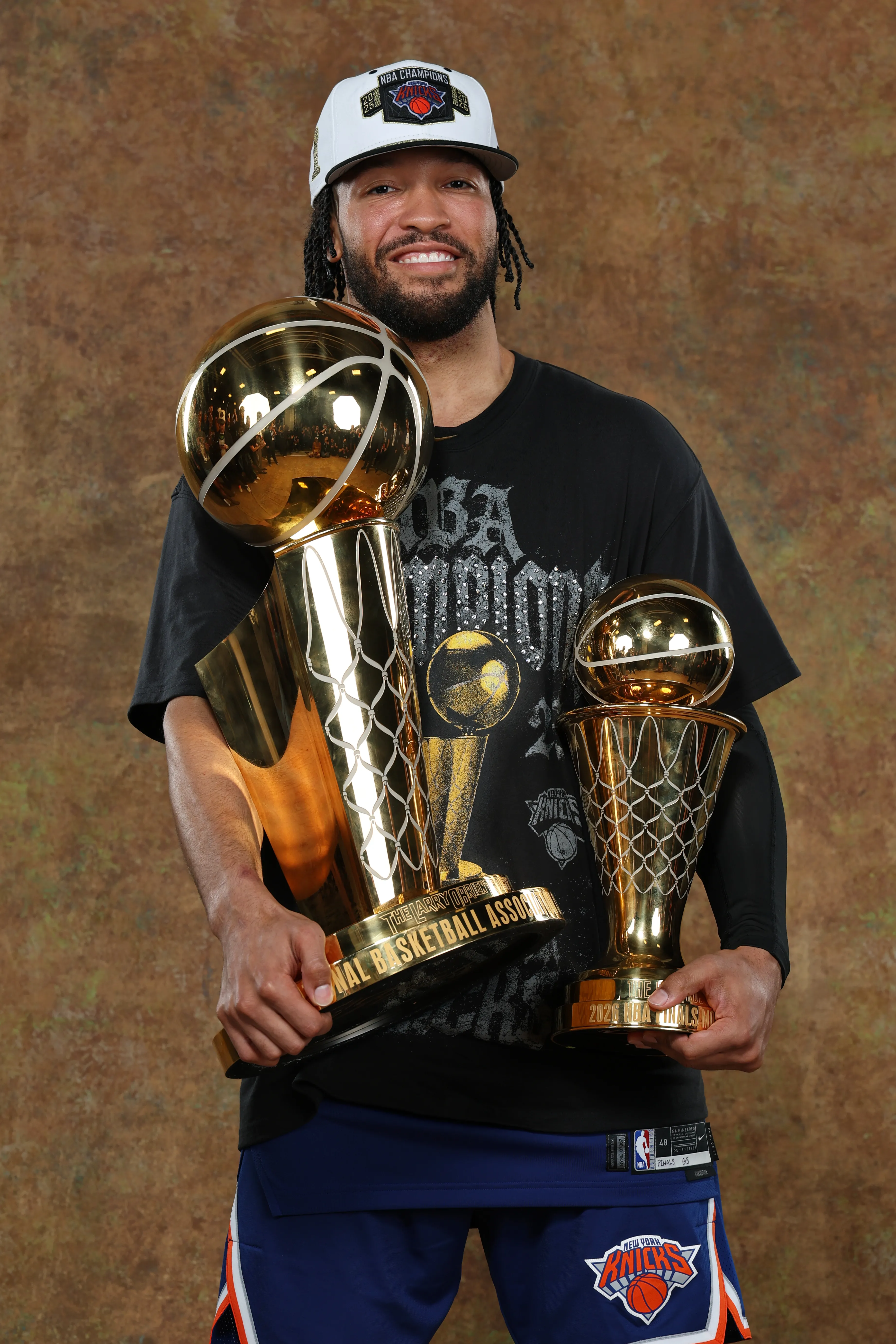 Jalen Brunson in a black shirt, blue shorts, and white "NBA Champions New York Knicks" cap, holding two golden NBA championship trophies.