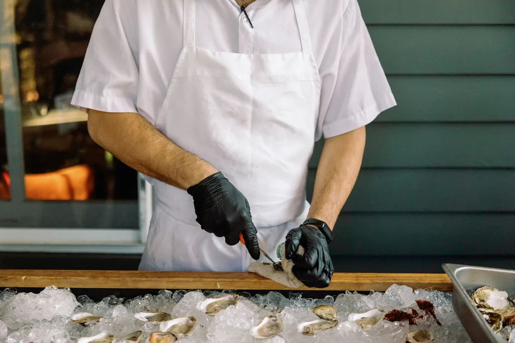 A caterer in an apron and black gloves shucking oysters at an oyster bar for a wedding.