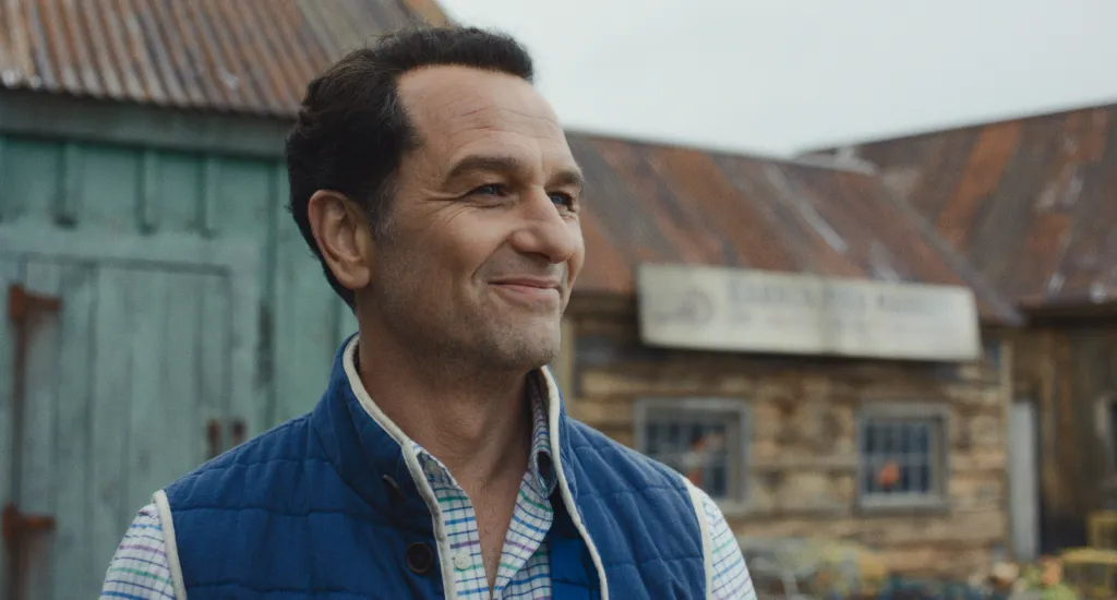 Matthew Rhys in Widow's Bay, wearing a blue quilted vest and a plaid shirt.