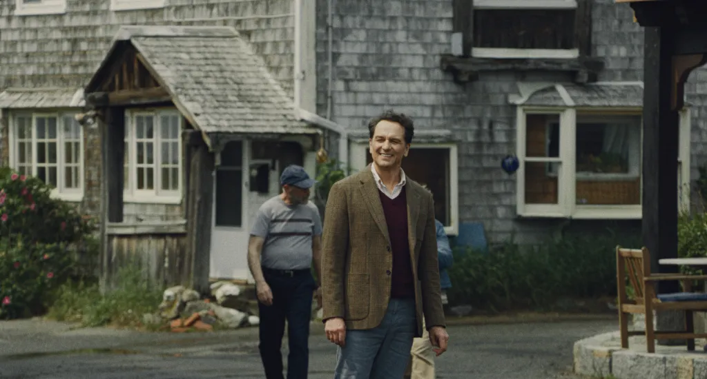 Matthew Rhys as a man in a brown tweed jacket, burgundy v-neck sweater, and collared shirt smiles and looks to his right.