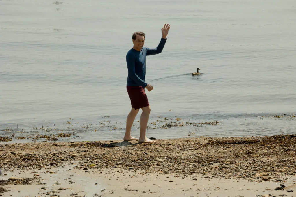 Matthew Rhys on a beach waving with a duck swimming nearby.