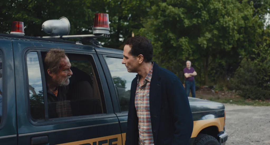 Matthew Rhys conversing with Stephen Root who is seated in a police car.