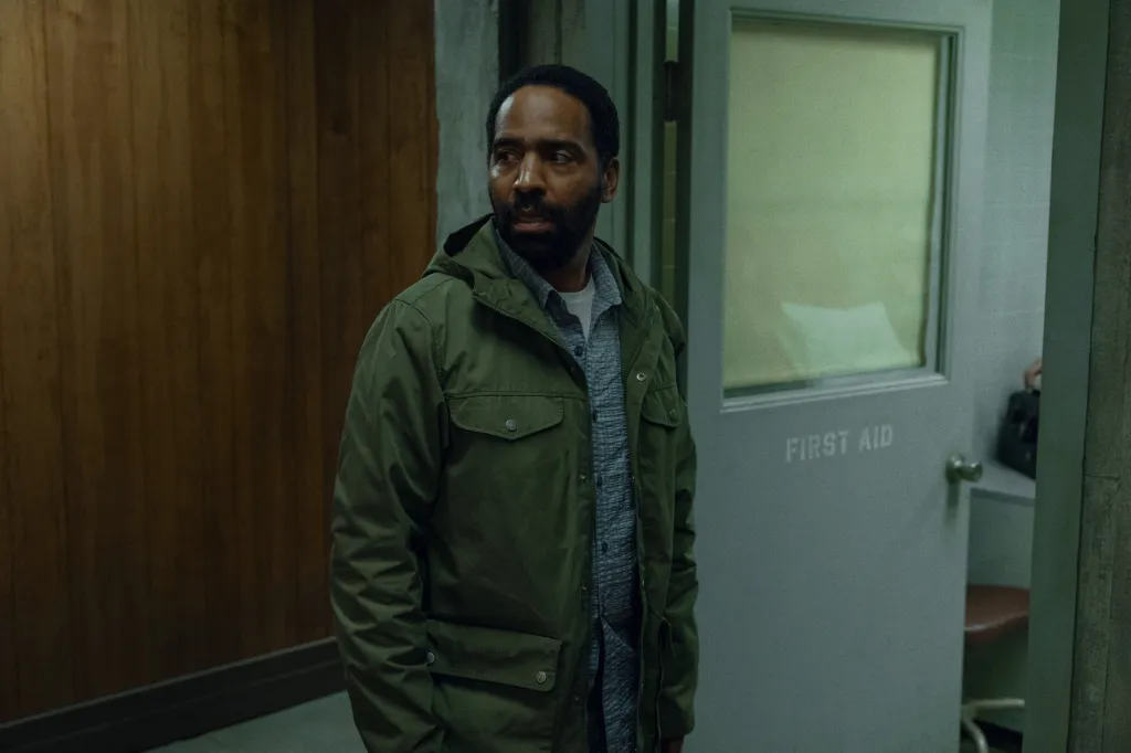 Man in an olive green jacket looks to the right of the frame, next to a "FIRST AID" door.