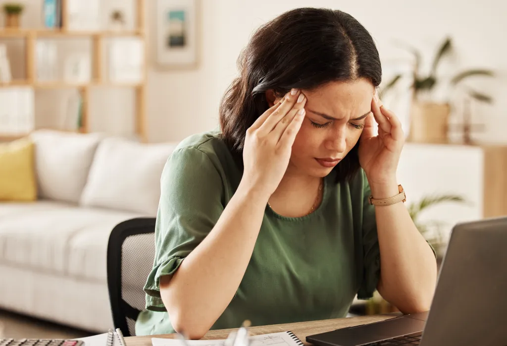 A woman with dark hair, wearing a green shirt, sits at a desk with a laptop, looking distressed and holding her head, suggesting a headache or stress from work.