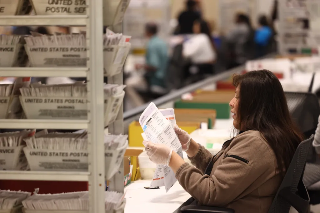 A worker in clear gloves sits at a desk, looking at an official ballot with multiple choices in a room with shelves of ballot boxes.