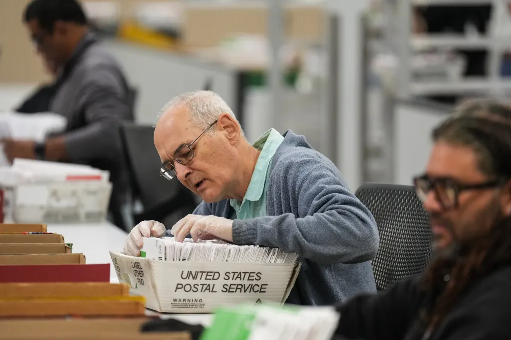 A worker wearing gloves sorts ballots in a United States Postal Service container.