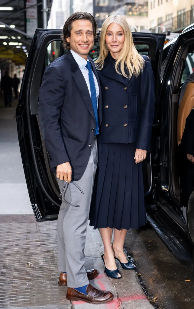 Writer/director Brad Falchuk, in a navy blazer, grey pants and a white shirt and blue tie, and actress Gwyneth Paltrow, in a blue jacket and blue skirt, standing next to an open car door.