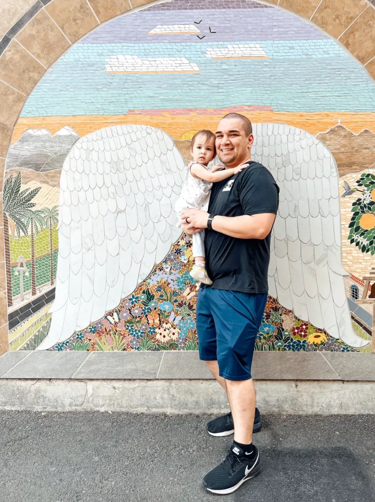 Lt. Colonel Gabriel Estrella holding his daughter in front of a mosaic mural of angel wings.