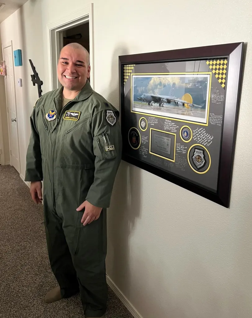 Lt. Colonel Gabriel Estrella in a flight suit stands beside a framed display including a photo of a B-52 bomber and patches.