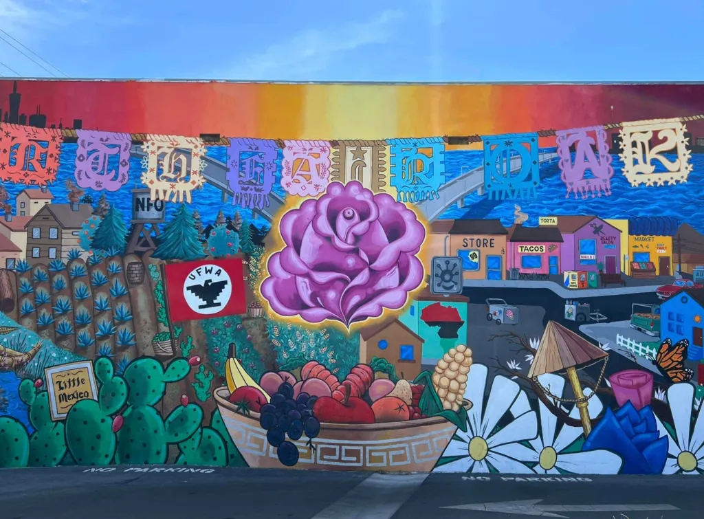 Illustration of a colorful mural depicting a vibrant community with houses, stores, fruits, flowers, and the letters "NFO CA" on papel picado banners, with a large purple rose at the center.