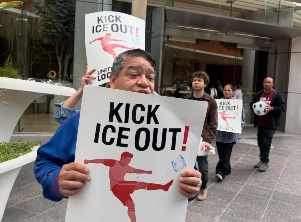 A person holds a "Kick ICE Out!" sign depicting a soccer player kicking an ice cube, while other protesters with similar signs are in the background.