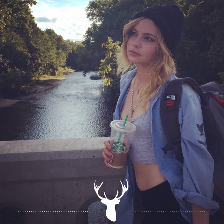 Daveigh Chase with a drink on a bridge over a river.