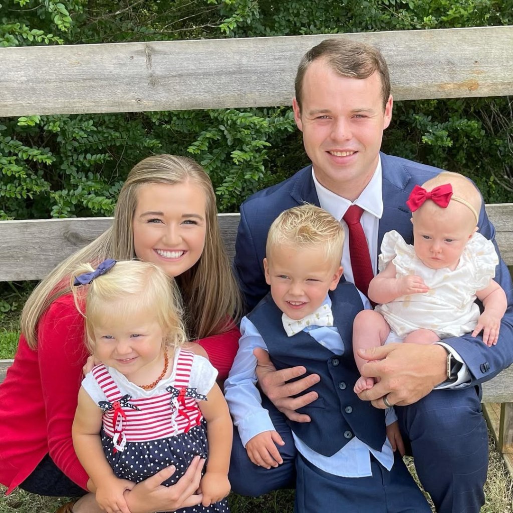 Joseph Duggar and Kendra Caldwell smiling with their three children: a blonde toddler girl, a blonde toddler boy, and a baby girl with a red bow in her hair.