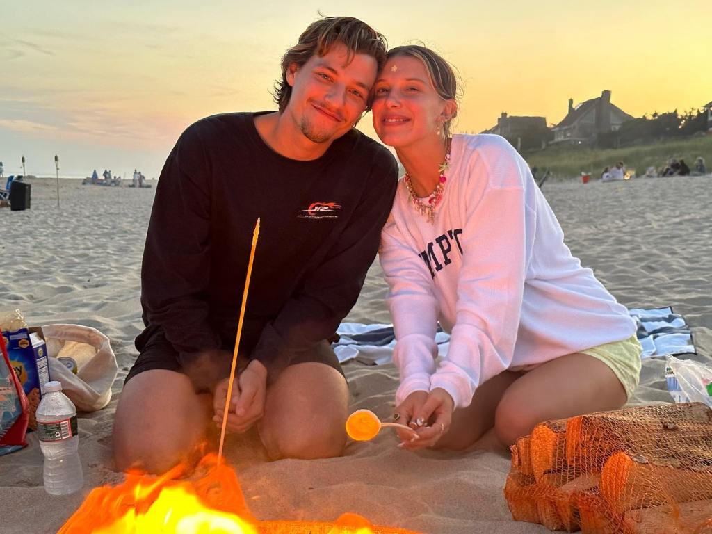 Millie Bobby Brown and Jake Bongiovi roasting marshmallows on a beach at sunset.