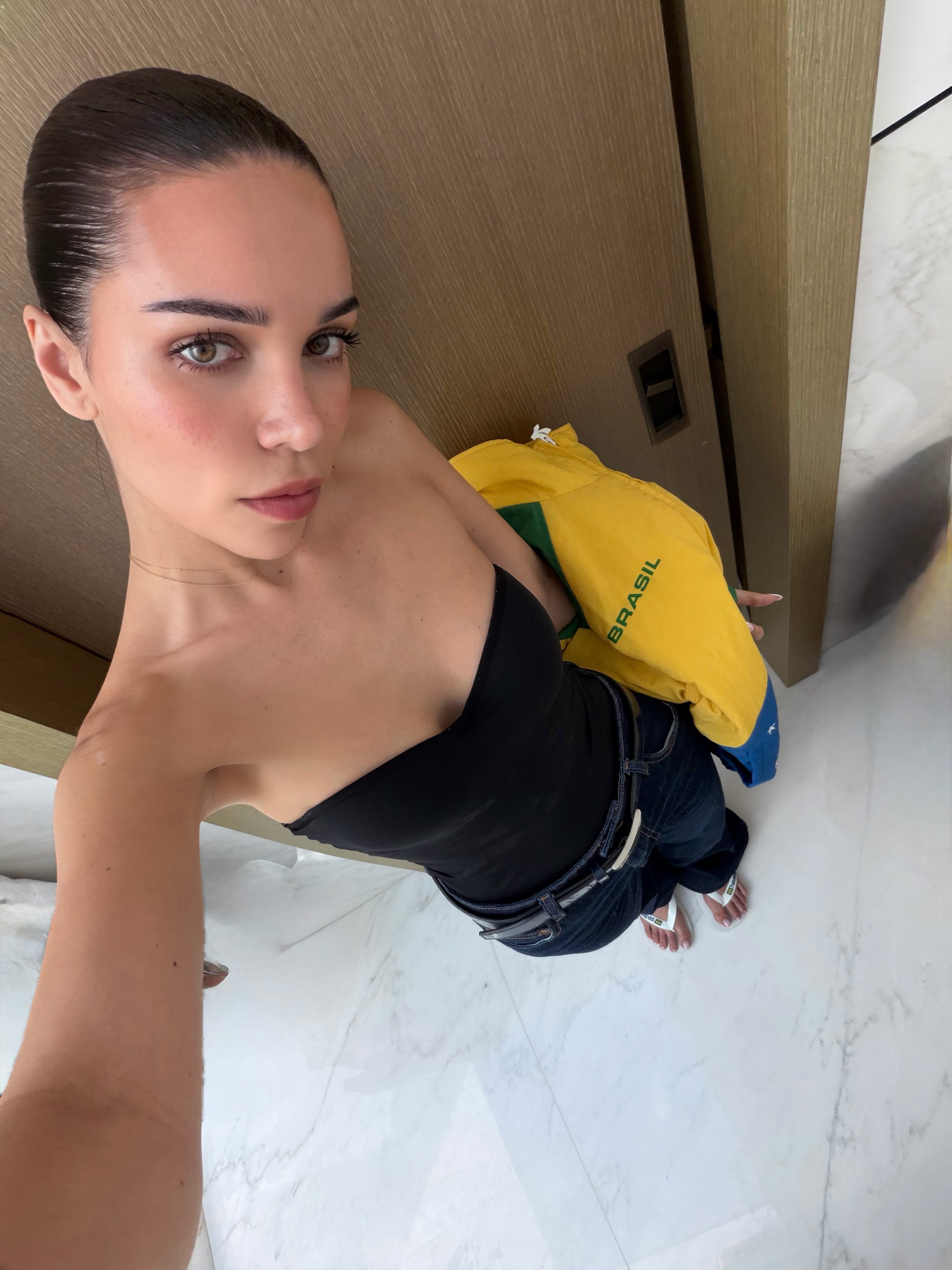 Selfie of Jackie Apostel wearing a strapless black top and dark jeans, holding a yellow jacket that says "BRASIL."