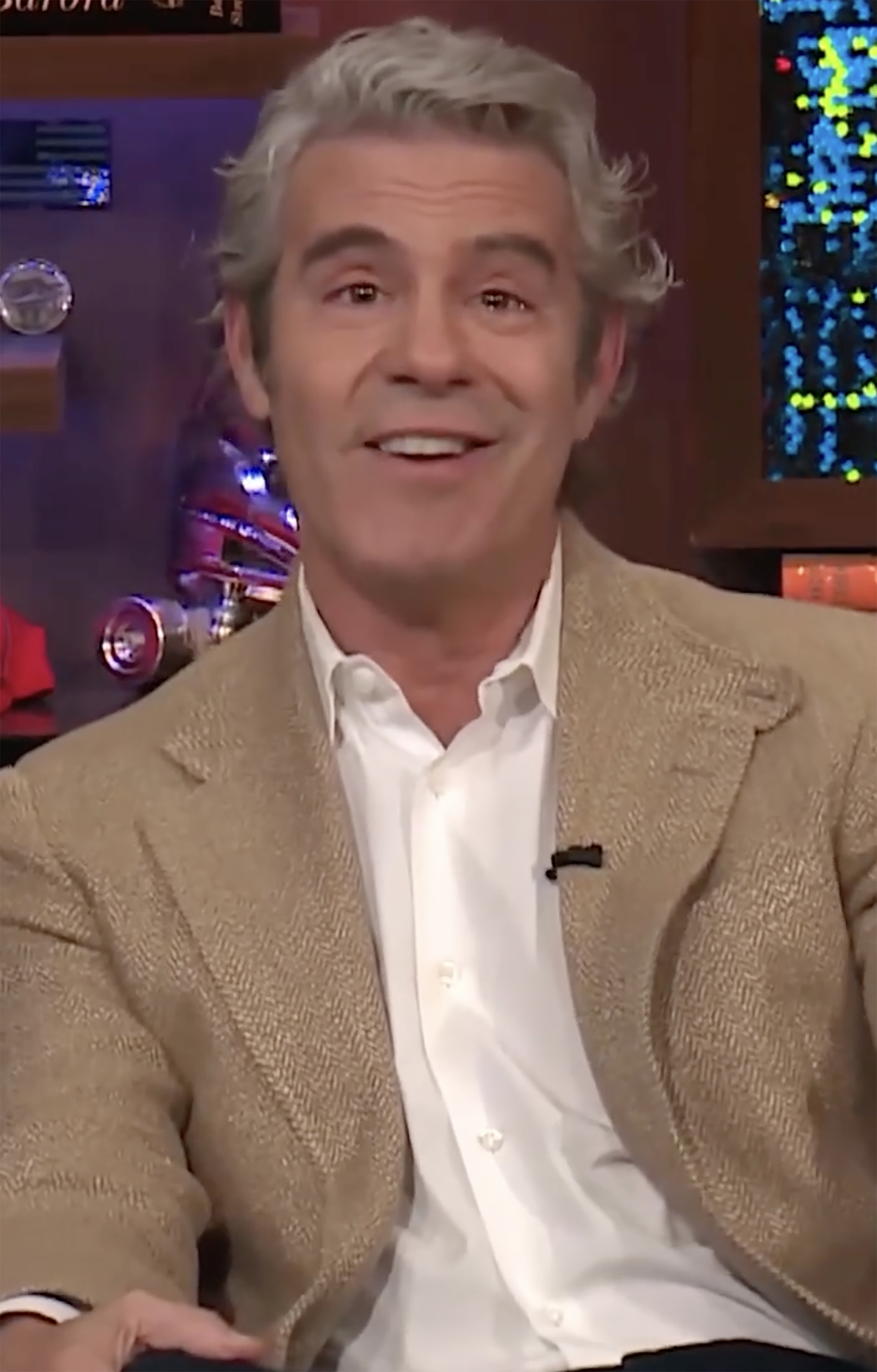 Andy Cohen appears tearful while wearing a white shirt and tan blazer.