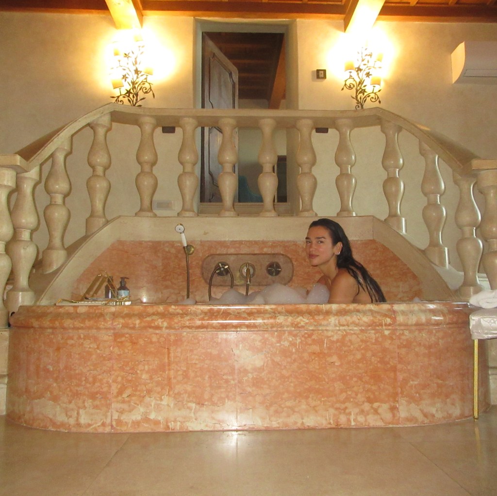 Dua Lipa in a bubble bath in a large, ornate marble tub.