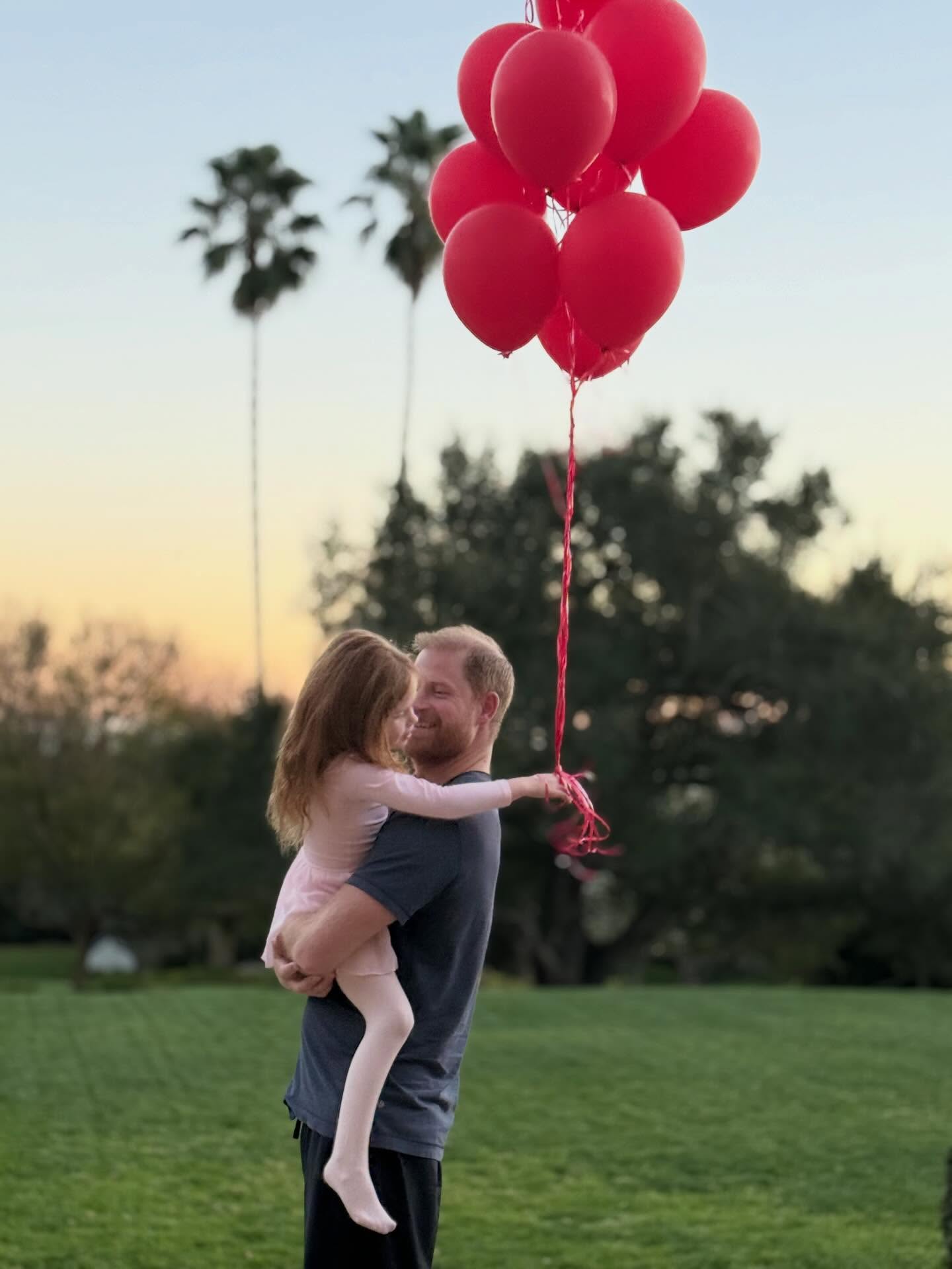 Prince Harry holding his daughter, Lilibet, who is holding red balloons.