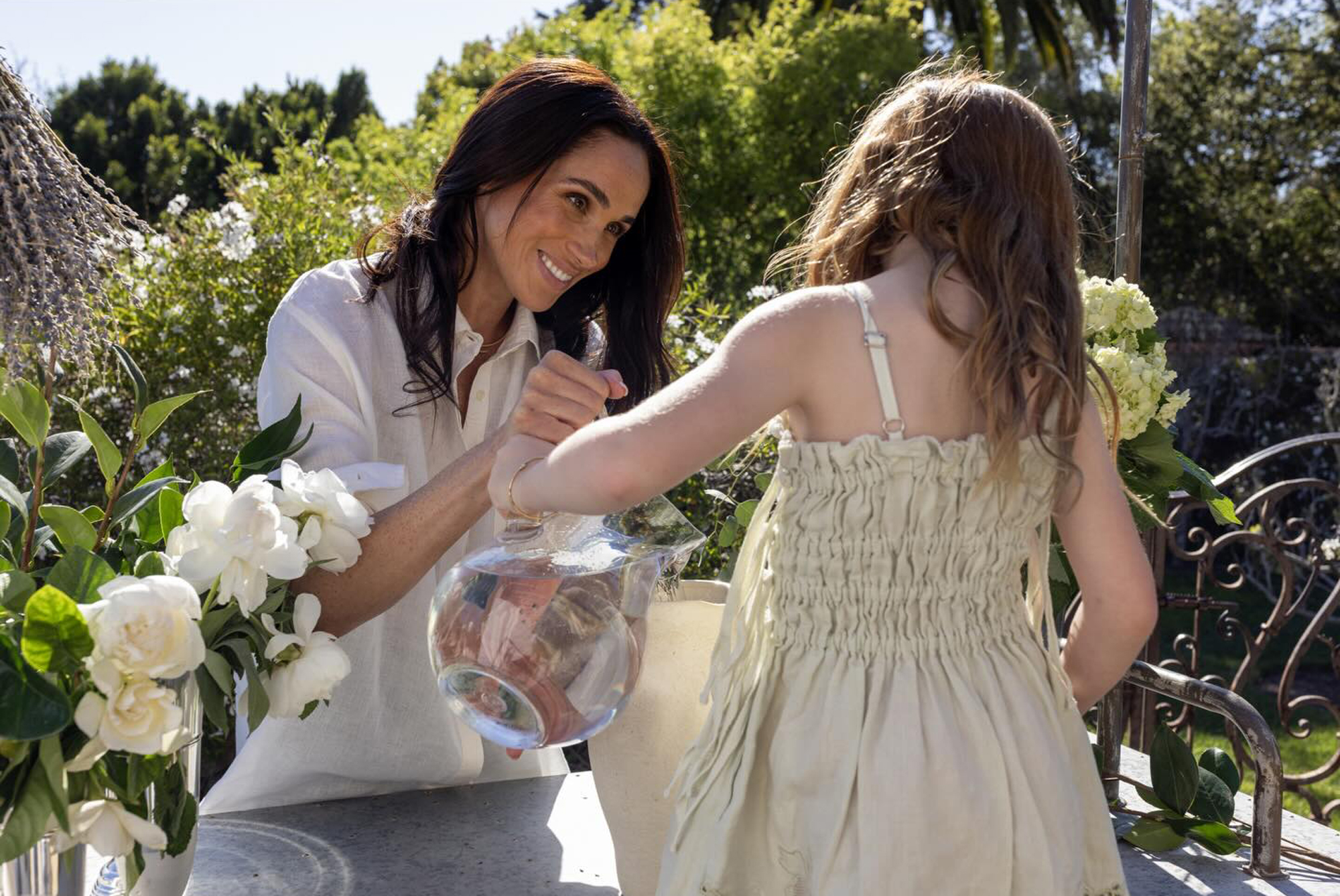 Meghan Markle smiles, helping Lilibet hold a glass pitcher of water.