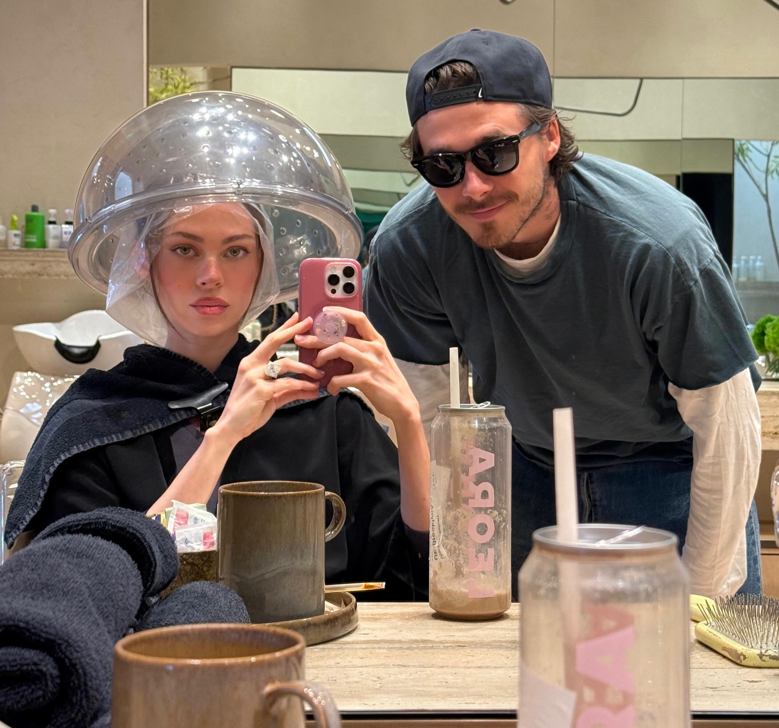 Nicola Peltz wearing a hair dryer hood while Brooklyn Beckham poses behind her.