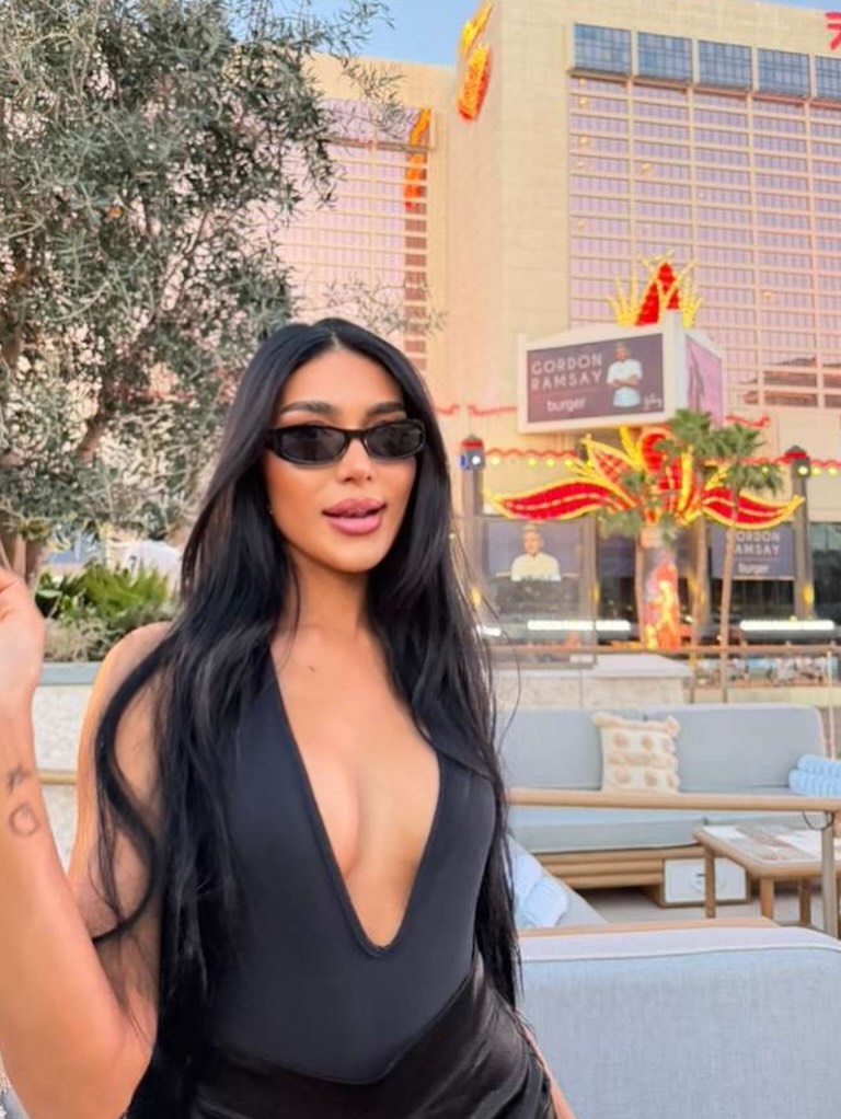 Thais Ramone wearing sunglasses and a black plunging top, with a hotel and "Gordon Ramsay Burger" sign in the background.