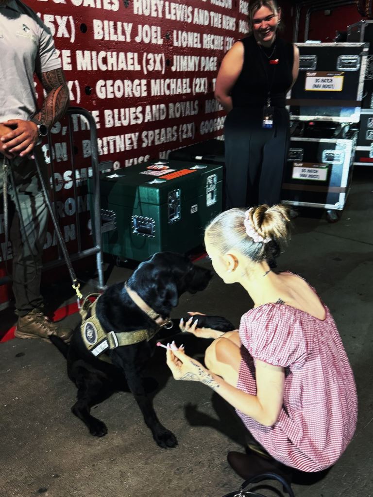 Ariana Grande crouching to pet a black dog wearing a service vest.