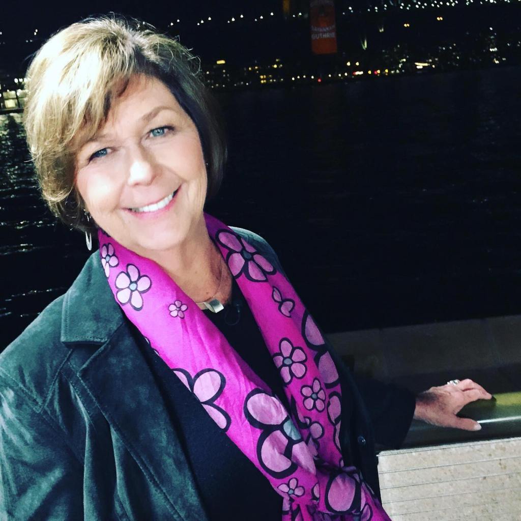 Nancy Guthrie smiles wearing a fuchsia scarf with flower patterns, with a city skyline at night in the background.
