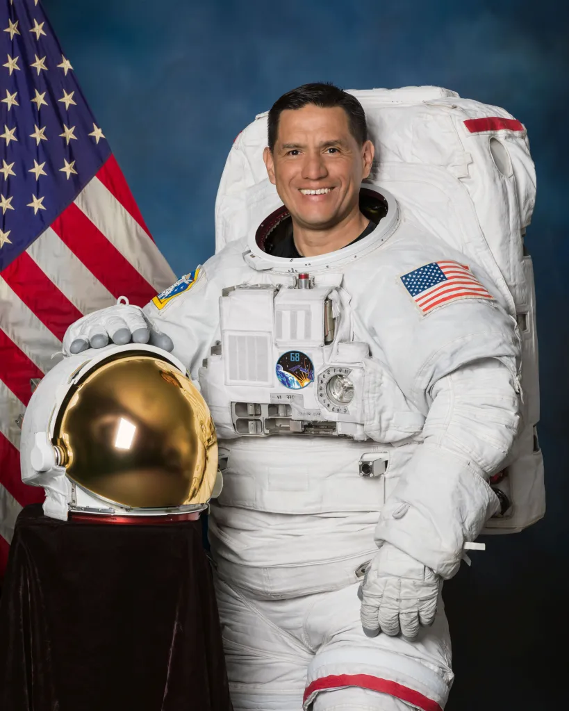 Astronaut Frank Rubio in a white spacesuit, holding a helmet, with the American flag in the background.