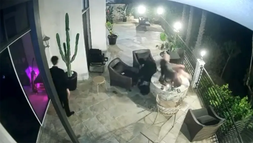 A man in a black jacket and pants pushes another man into a fire pit on a patio at night.
