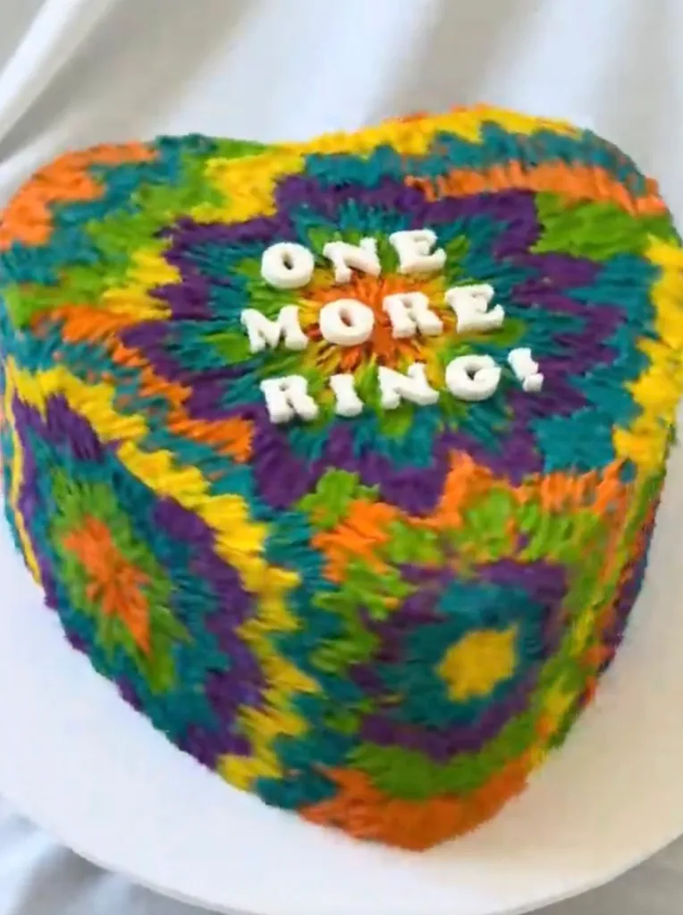 A heart-shaped cake with tie-dye frosting that says "ONE MORE RING!"