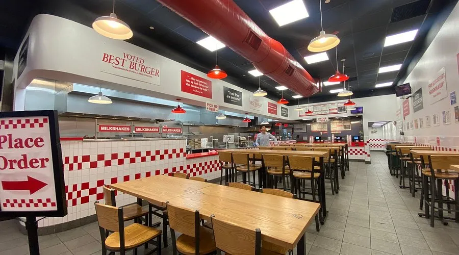 Interior of a Five Guys restaurant with red and white checkered walls, wooden tables, and a "Place Order" sign.