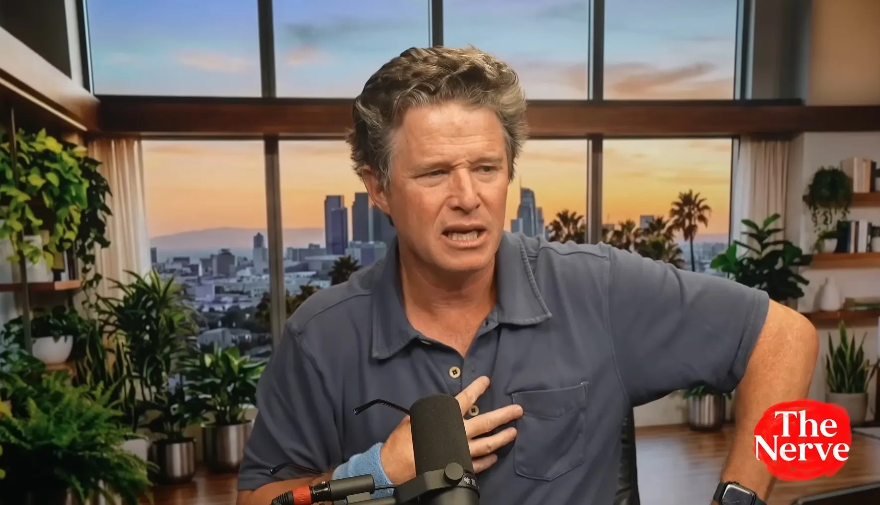 Billy Bush in a blue polo shirt gestures while speaking into a microphone.