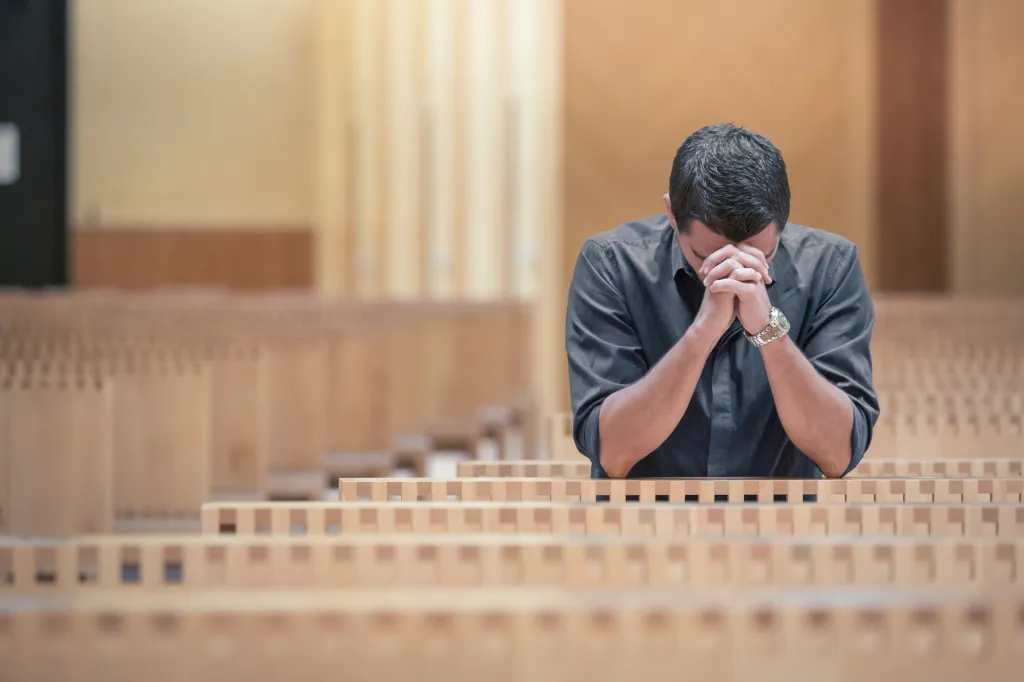 Young man with a beard, wearing a blue shirt, praying in a modern church with his hands clasped together.
