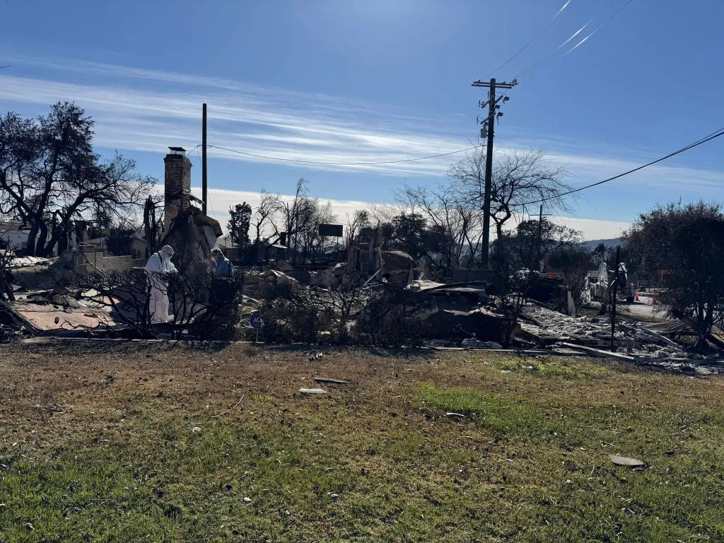 A residential area destroyed by fire, showing rubble, a chimney, and a charred landscape under a blue sky, with two people in the debris.