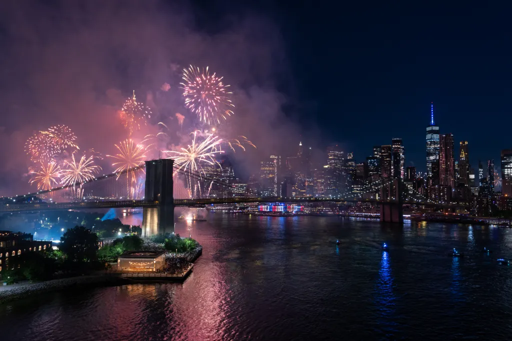 Fireworks illuminating the sky above the Brooklyn Bridge and New York City skyline.