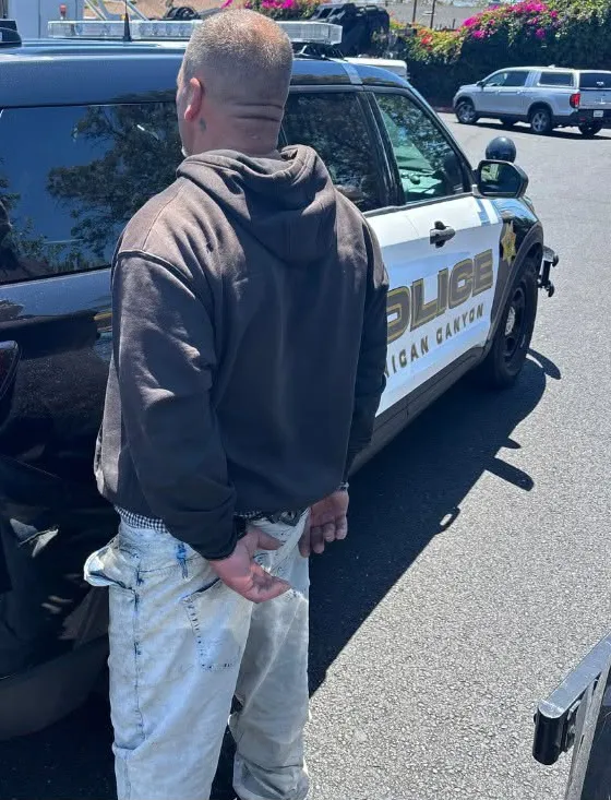 A man in handcuffs stands facing away from the camera, next to an American Canyon Police Department vehicle.