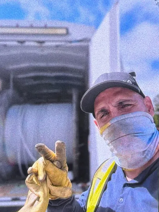 A man wearing a face covering, yellow safety vest, and work gloves gives a peace sign in front of a white truck.