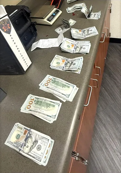 Hundreds of US dollar bills are laid out on a counter next to a police logbook and a scale.