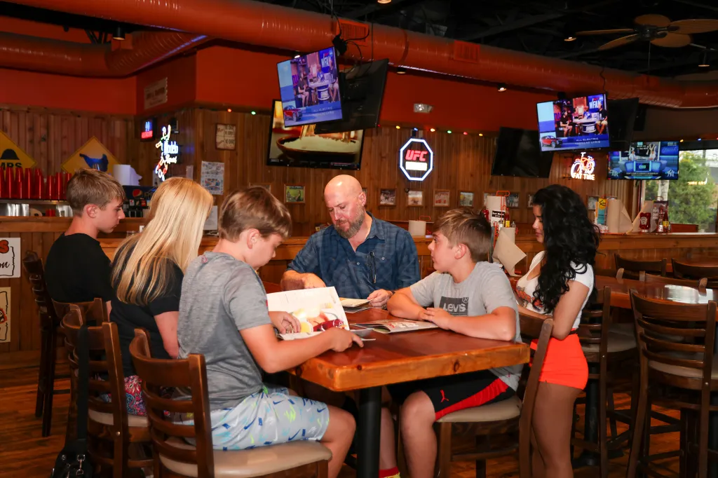 The Llewellyn family, including a father, two sons, a blonde woman, and a Hooters waitress, sitting at a table in a Hooters restaurant in Farmingdale, NY.