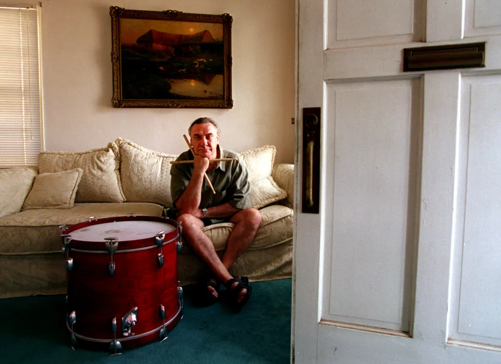 Bill Ward, former drummer of Black Sabbath, sits on a couch in his home with drumsticks and a drum.