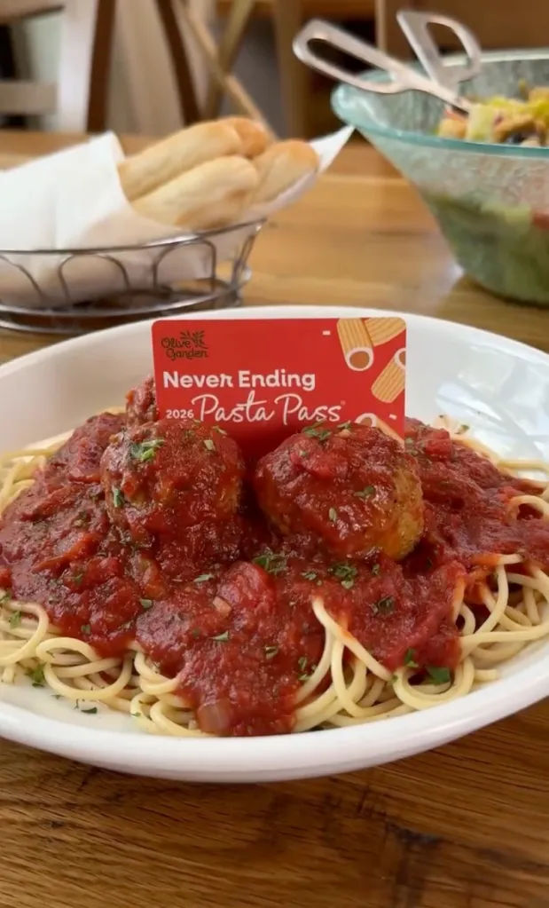 An Olive Garden Never Ending Pasta Pass stands upright in a plate of spaghetti with meatballs, sauce, and parsley.