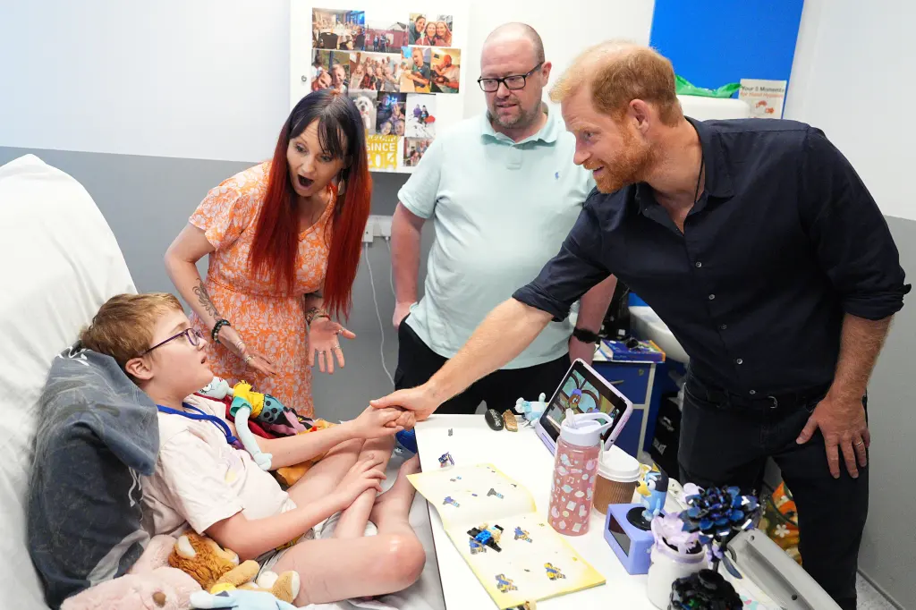 Prince Harry, in a navy blue shirt and black jeans, holds hands with patient Alec Hill, in a cream T-shirt, as his parents Kelly and Stuart Hill look on.