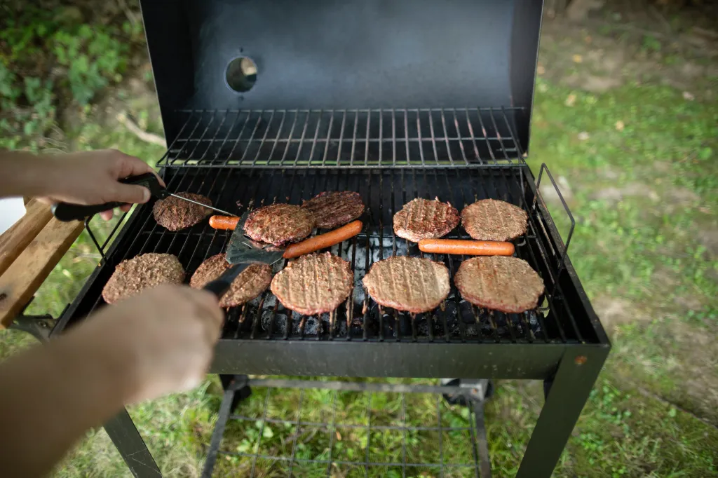 Person's hands checking meat temperature of burgers and flipping hot dogs on a backyard grill.