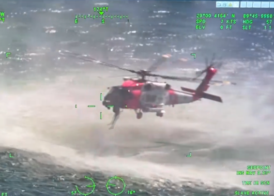 A red and white helicopter hovering over a turbulent body of water, viewed from a slightly higher angle with green digital flight data overlaying the image.
